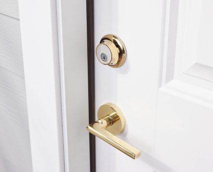 Polished brass finish on front door knob and deadbolt, shining in the light. Shown on white door with white trim and grey siding.